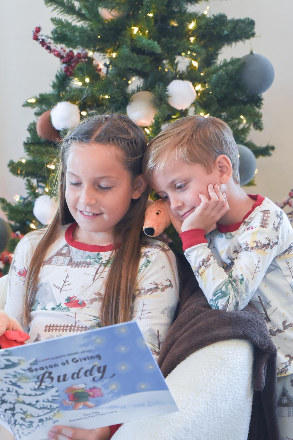 Two children in pajamas sitting in front of a decorated Christmas tree, reading Season of Giving Buddy with Buddy plush.